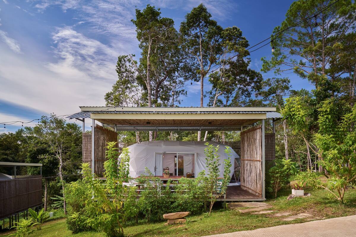 A covered outdoor structure with a large tent-like space, surrounded by lush greenery and trees.