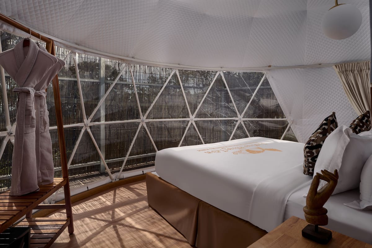 A room with a bed, a wooden bench, a robe rack, and a large window with a view of trees.