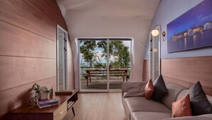 A modern living room with a wooden floor, a sofa with cushions, a small table with a book and a decorative item, and a large window with a view of a balcony.