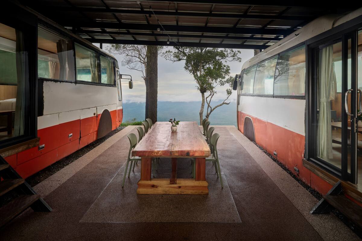 A covered outdoor dining area with a wooden table and chairs, surrounded by large windows and a view of the mountains.