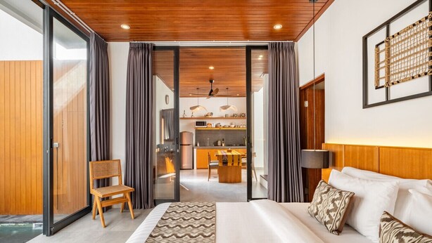 Quarto de hotel moderno com uma cama grande, cabeceira de madeira, área de jantar e cozinha ao fundo, e porta de vidro deslizante que dá para uma área externa.