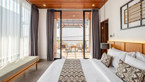 Quarto de hotel moderno com uma cama grande, um banco, uma varanda com cadeiras e vista para o exterior.