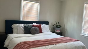 Un dormitorio con cama, ventilador de techo, ventana con persianas, planta en la mesita de noche y mesita de noche.
