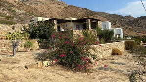 A Mediterranean-style villa with stone walls and a flat roof, surrounded by dry vegetation and mountains.