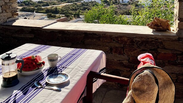 A table set with a striped tablecloth, a bowl of watermelon, a coffee pot, and a straw hat.