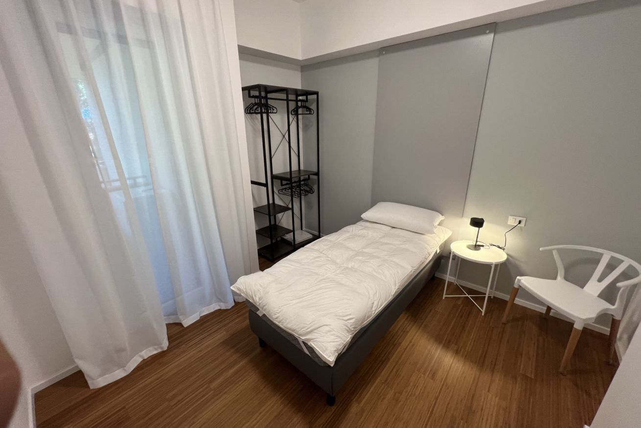 A bedroom with a bed, a bedside table with a lamp, a chair, and a clothing rack.