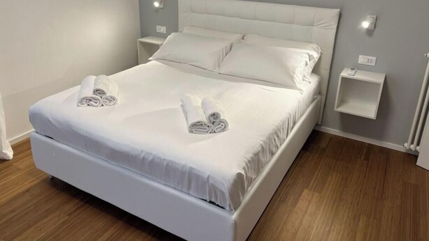 A neatly made bed with white linens and towels, a padded headboard, bedside tables, and a light grey wall. - Villa Riviera 287 - Appartamento Mare (Pescara)
