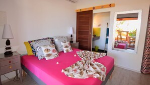 A bedroom with a pink bedspread, multiple pillows with a nature print, a bedside table with a lamp, and a view of an outdoor area through a window. - Direct Privilege Rate - 2 adults (Saint-Paul)