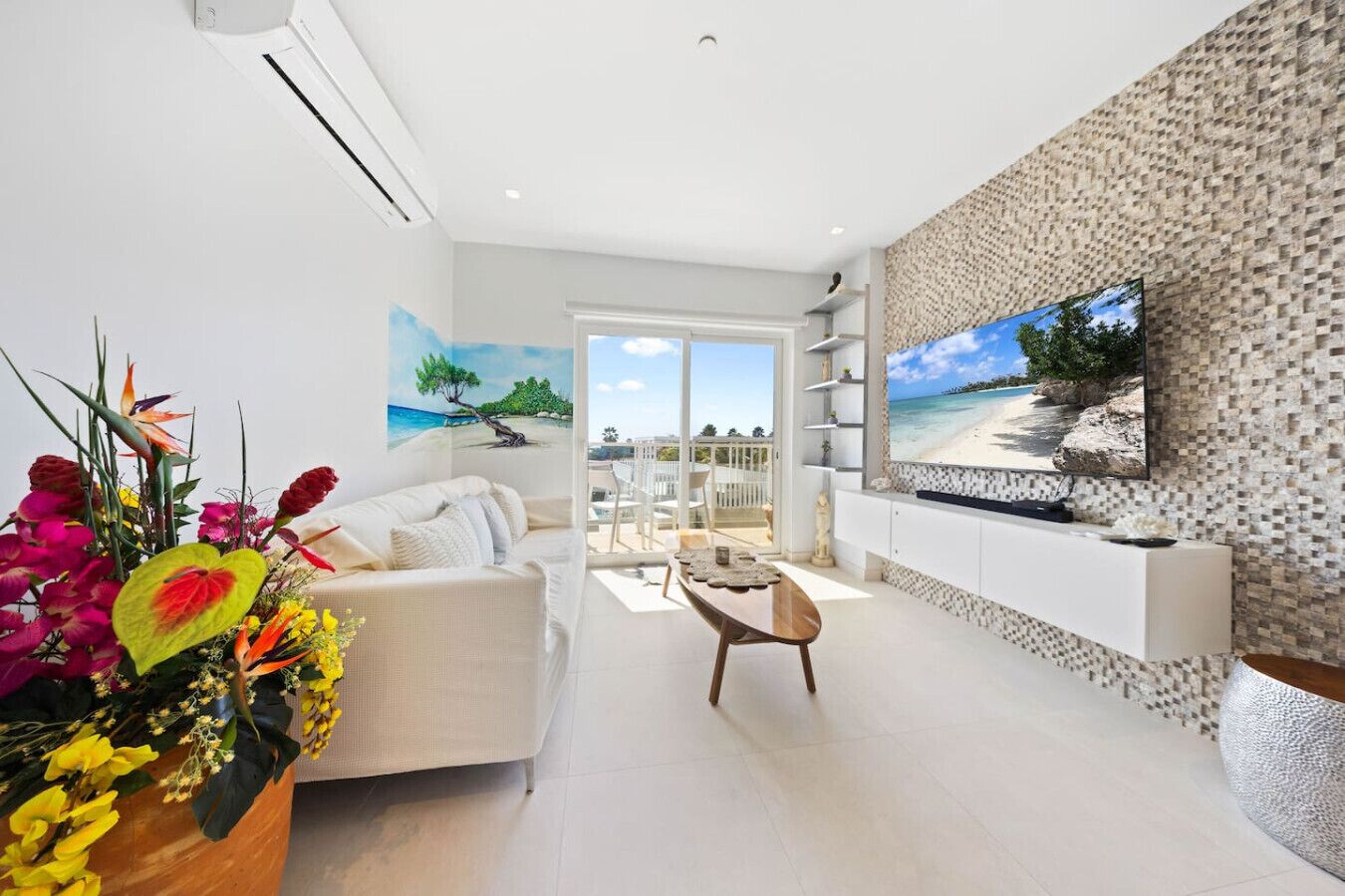 A modern living room with a sofa, coffee table, and a TV mounted on a textured wall. There is a balcony with a view of the beach.