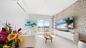 Uma sala de estar moderna com sofá, mesa de centro e TV fixada em uma parede texturizada. Há uma varanda com vista para a praia.