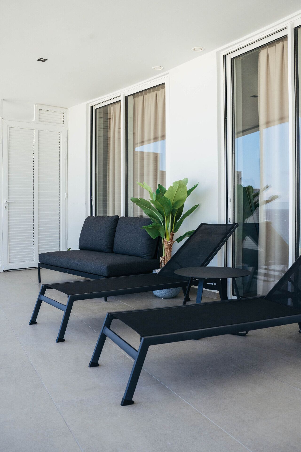 A modern outdoor seating area with a sofa and two lounge chairs, a potted plant, and a glass door leading to a balcony.
