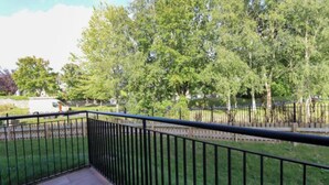 A balcony with a black metal railing, a paved floor, and a view of a green park with trees and a white vehicle in the background. - Charming 2-Bed Apartment in Dublin's Countryside (Ballyboughal)
