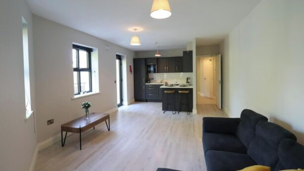 A spacious living room with a dark sofa and a wooden coffee table, a kitchen with dark cabinets and a bar area, and a large window providing natural light. - Charming 2-Bed Apartment in Dublin's Countryside (Ballyboughal)