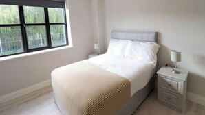 A bedroom with a bed, a nightstand, a lamp, and a window with a view of greenery. - Charming 2-Bed Apartment in Dublin's Countryside (Ballyboughal)