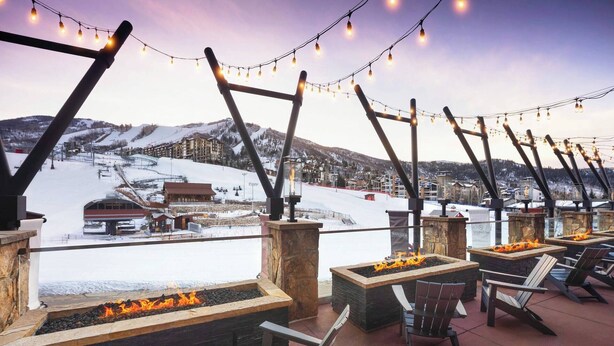 An outdoor seating area with fire pits and chairs, overlooking a snowy ski slope.