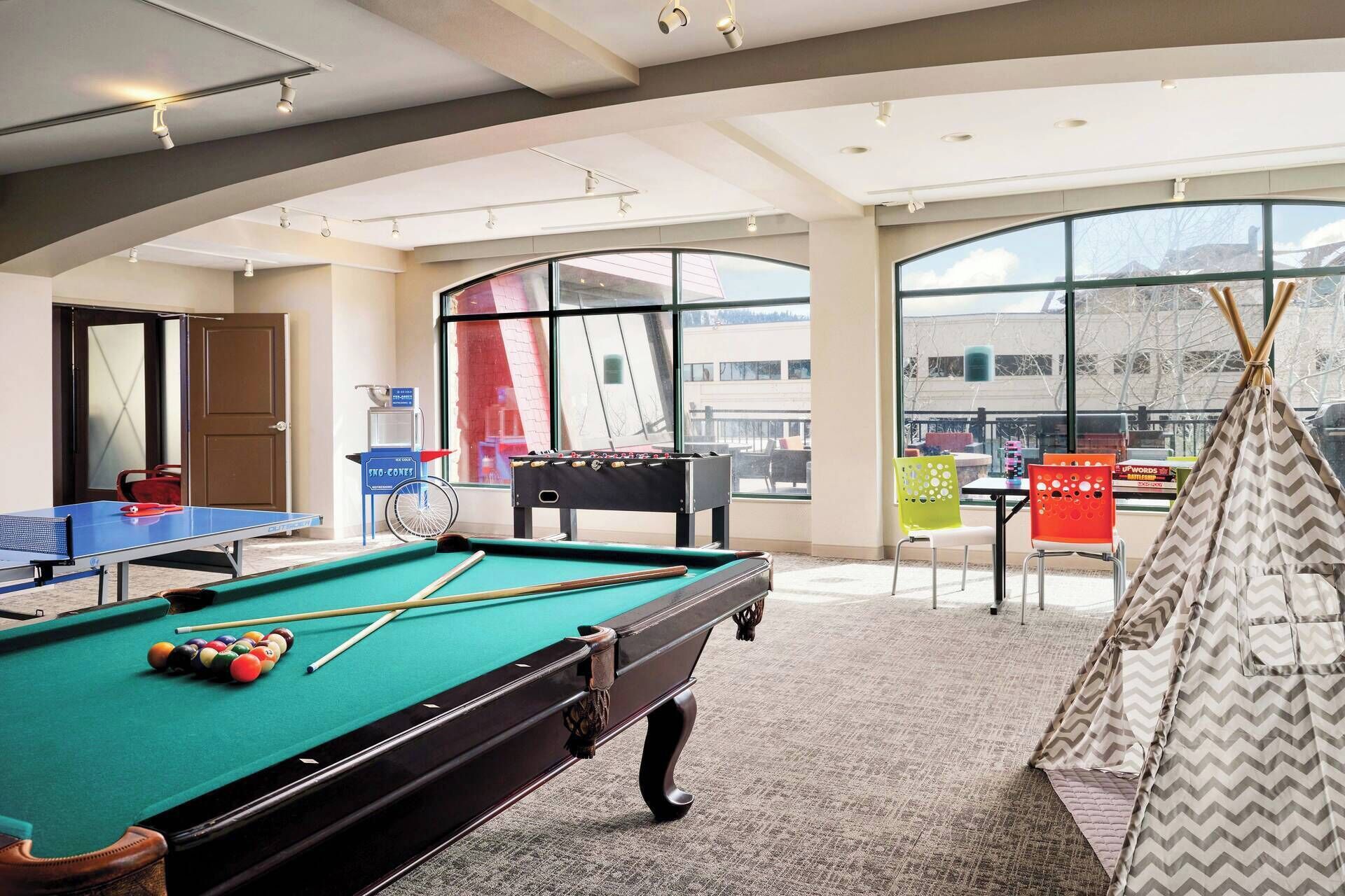 A pool table, foosball table, and a teepee in a room with large windows and a view of the outside.