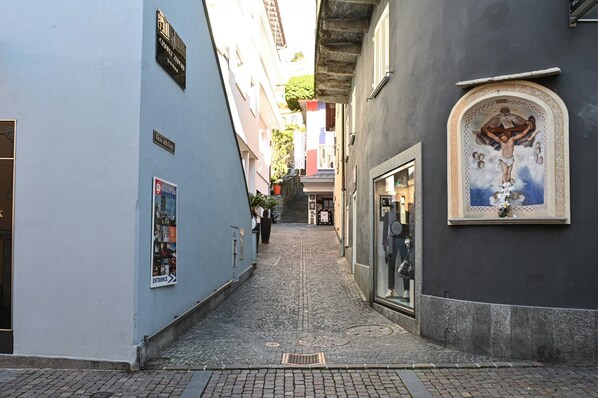 A narrow cobblestone alley with buildings on either side, a mural of a crucifixion on the right wall, and a sign indicating an entrance to a building on the left.