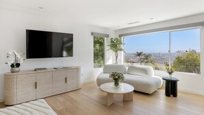 A modern living room with a flat-screen TV, a sofa, a coffee table, and a large window offering a city view. - Hollywood Hills Retreat • Pool + Spa + Outdoor Dining + Views | Skyline Chalet (Los Angeles)