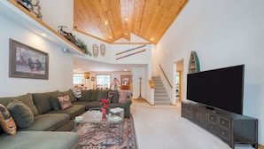 A spacious living room with a large flat-screen TV, a green sofa, a coffee table, and a staircase leading to an upper level. - Pinewood Lodge by AvantStay | Cozy Cabin Near Ski Resorts & Hiking (Truckee)