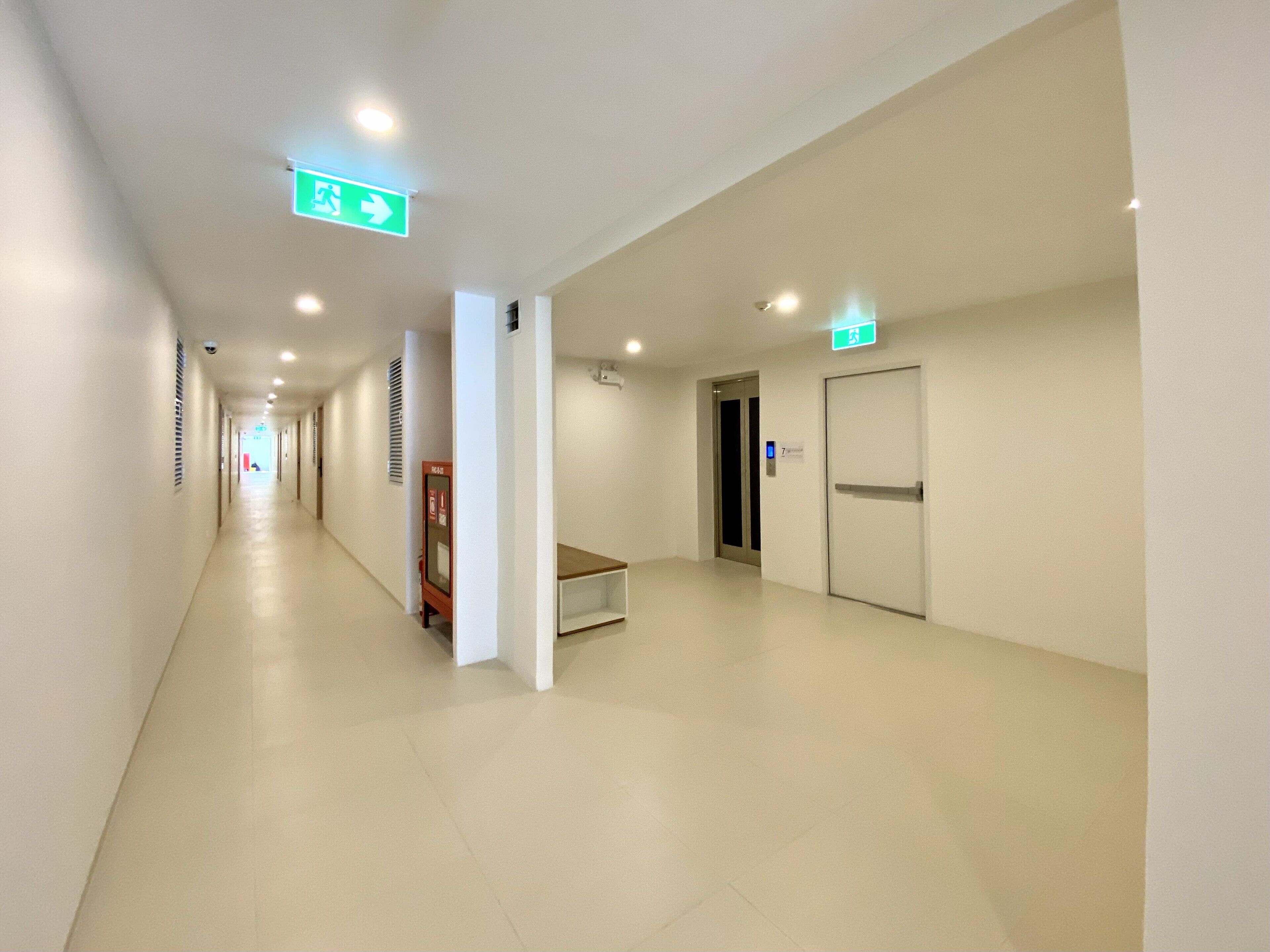 A long, narrow hallway with white walls and a tiled floor, leading to multiple doors and an emergency exit sign.