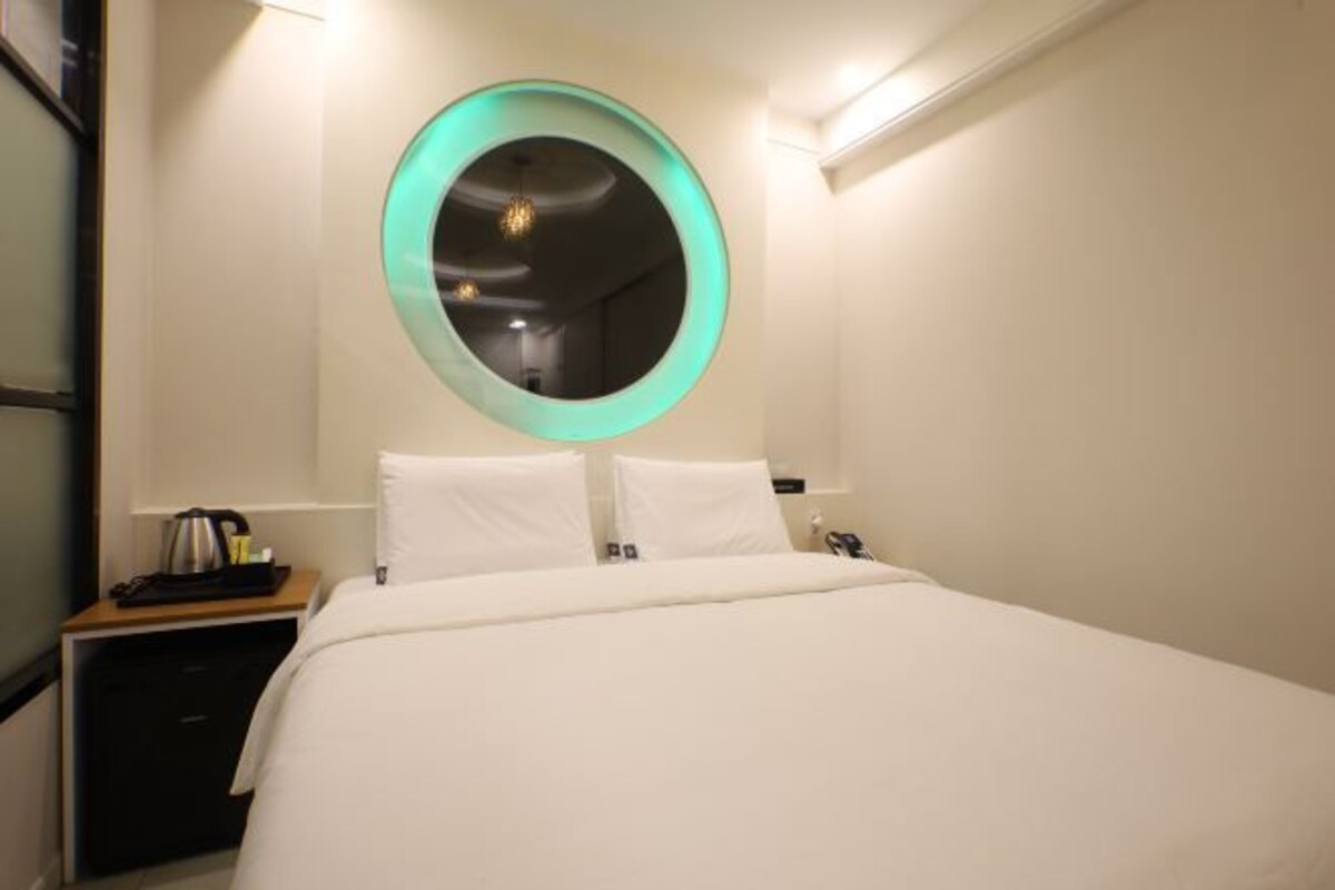 A hotel room with a bed, a round mirror with a teal frame, a bedside table with a kettle, and a nightstand.