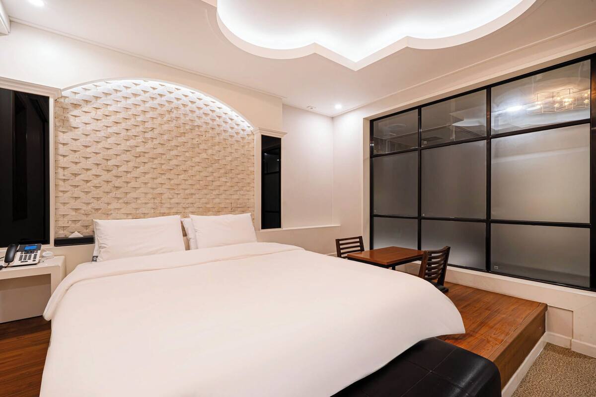 Une chambre d’hôtel moderne dotée d’un grand lit, d’un bureau avec un téléphone, d’une chaise et d’une grande fenêtre avec des panneaux de verre dépoli.