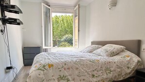 A bedroom with a bed, a nightstand, a dresser, and an open window with a view of greenery.
