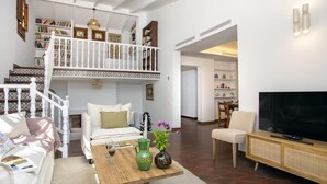 Een ruime woonkamer met een hoge plafond, een houten trap met witte leuningen, een flatscreen televisie en een eethoek met een tafel en stoelen.