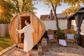 A sauna with a wooden exterior, a person entering, and two wooden chairs with cushions.