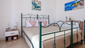 Ein Schlafzimmer mit einem Bettgestell aus grünem Metall, zwei herzförmigen Kissen, einem kleinen Nachttisch mit Lampe, einem Beistelltisch und einem Wandbild eines blauen Vogels.