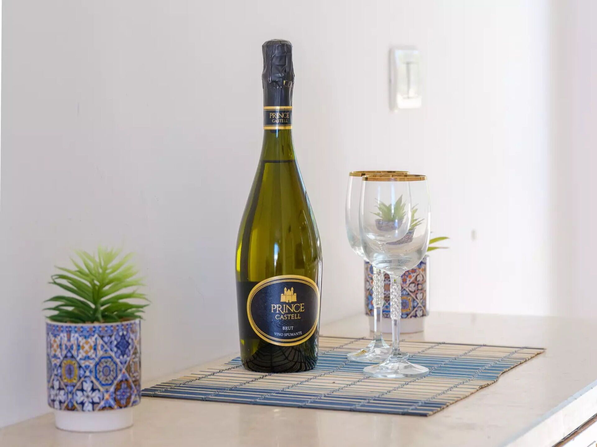En flaska av vinet Prince Castell Brut står på ett bord tillsammans med en dekorativ bordsduk, ett glas och en krukväxt.