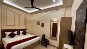A hotel room with a large bed, a ceiling fan, a TV, and a small desk. - The Commune ISKCON (Varanasi)