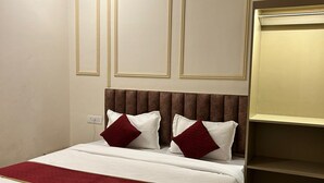 A neatly made bed with a red and white bedspread, a brown headboard, and two red pillows. There is a small bedside table with a lamp, and a wardrobe with a mirror. - The Commune ISKCON (Varanasi)