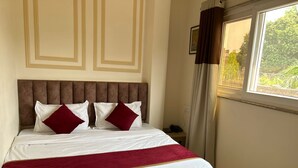 A neatly made bed with white linens and a red throw, a brown upholstered headboard, and a window with curtains. - The Commune ISKCON (Varanasi)