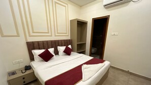 A neatly made bed with white and maroon bedding, a wooden headboard, and a red throw blanket. - The Commune ISKCON (Varanasi)