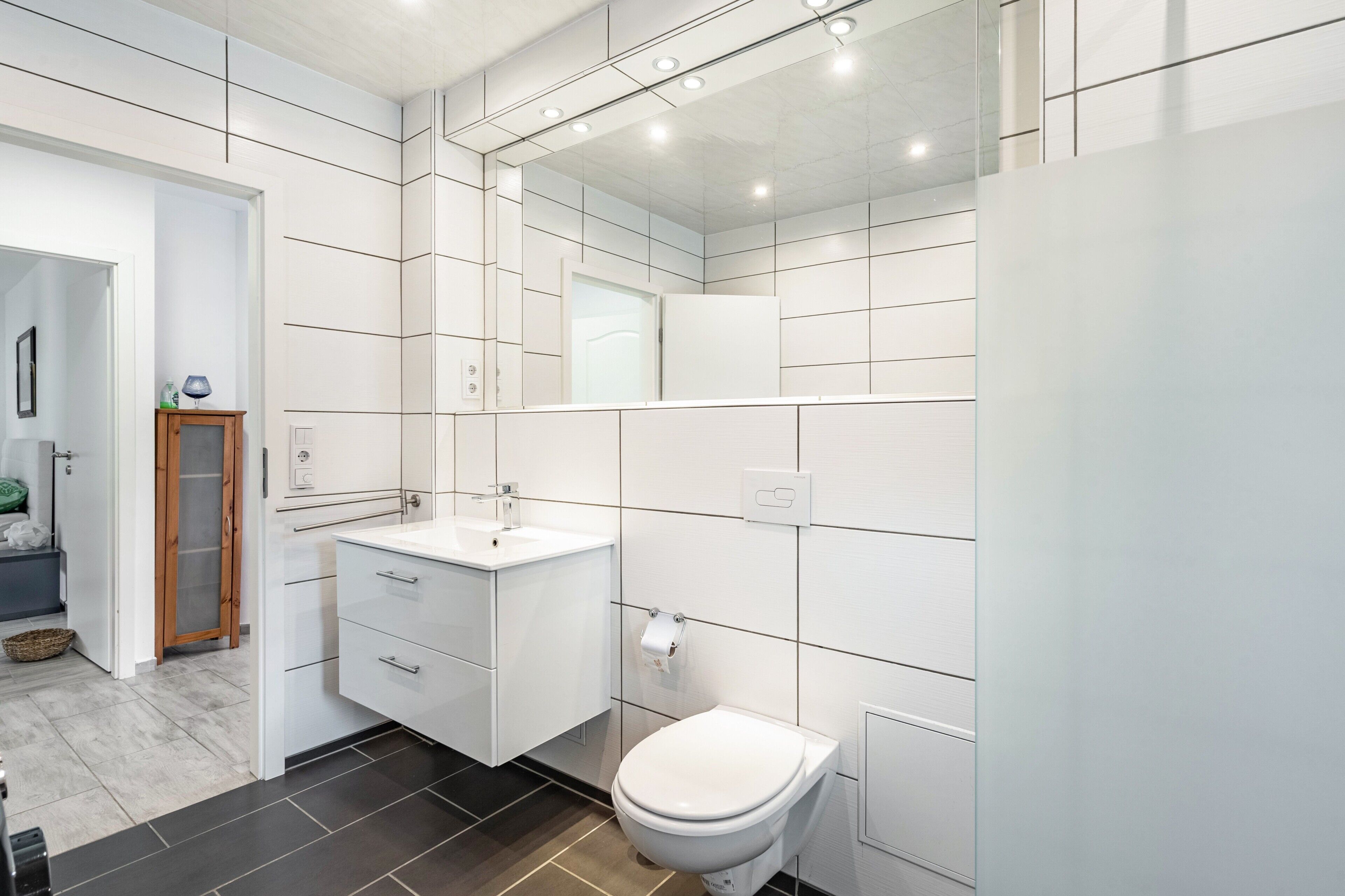 Un baño moderno con azulejos blancos, un lavamanos blanco y un inodoro.