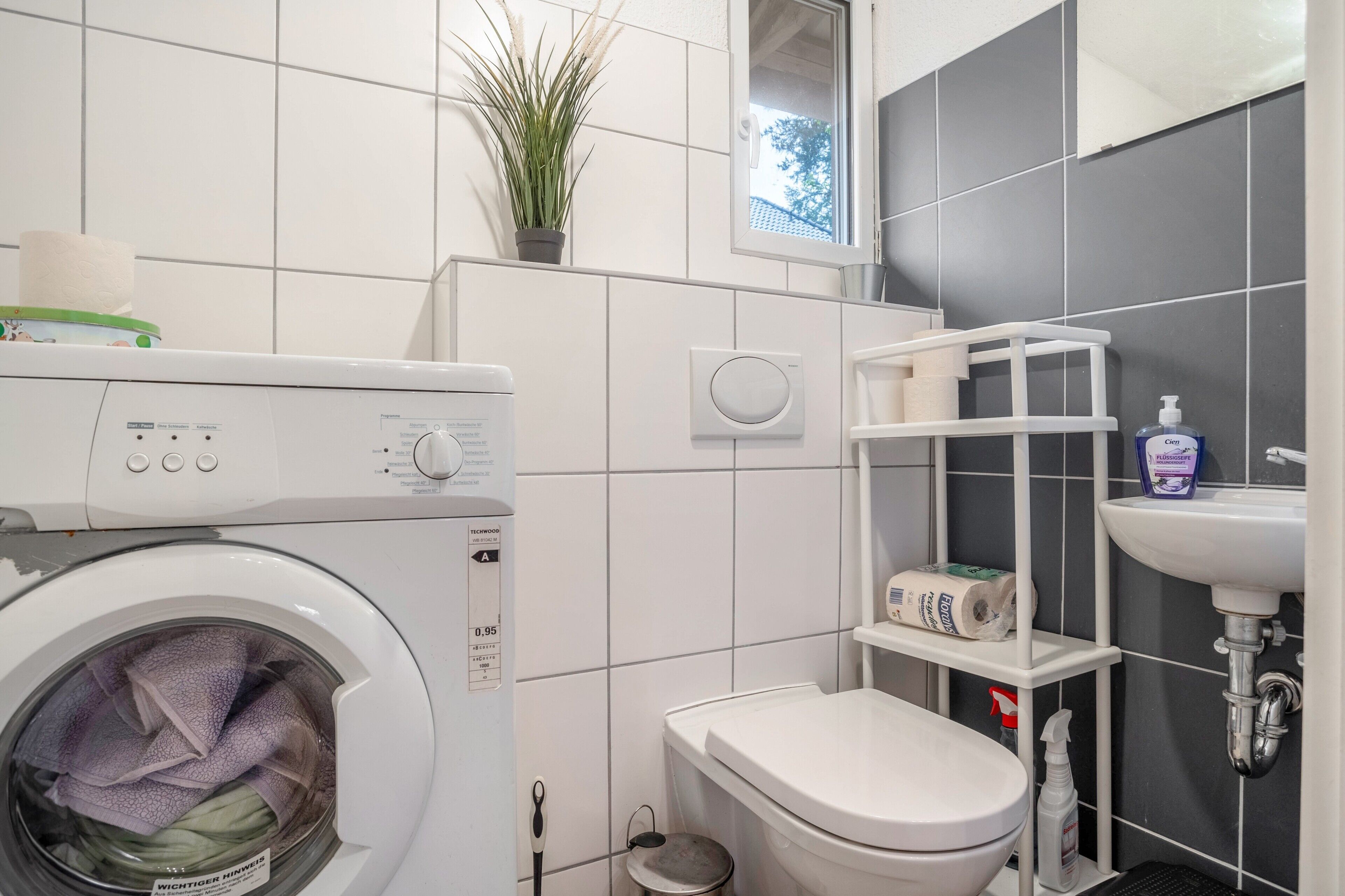 A compact bathroom with a washing machine, toilet, sink, and a small shelf with toiletries.