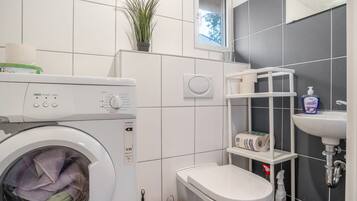 A compact bathroom with a washing machine, toilet, sink, and a small shelf with toiletries.