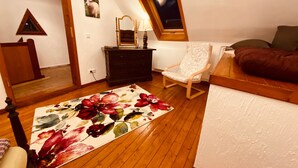 Une pièce avec un plancher en bois, un tapis à motifs floraux, un fauteuil à bascule, un lit, une commode, un miroir et un tableau représentant un mouton accroché au mur.