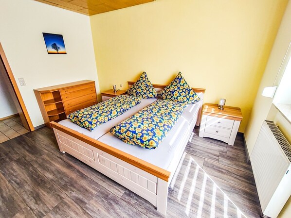 Un lit double avec une literie à motifs floraux, une tête de lit en bois, une table de chevet avec une lampe et un radiateur.