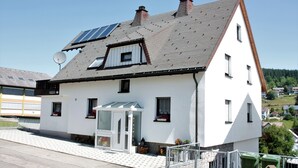 A white building with a sloped roof, solar panels, and a small porch with a glass door. - Apartment '45qm' with Mountain View, Shared Garden and Wi-Fi (Schonach im Schwarzwald)