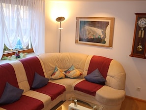 Un salon avec un canapé beige, des coussins rouges et bleus, un tableau encadré, une lampe sur pied et une horloge au mur.
