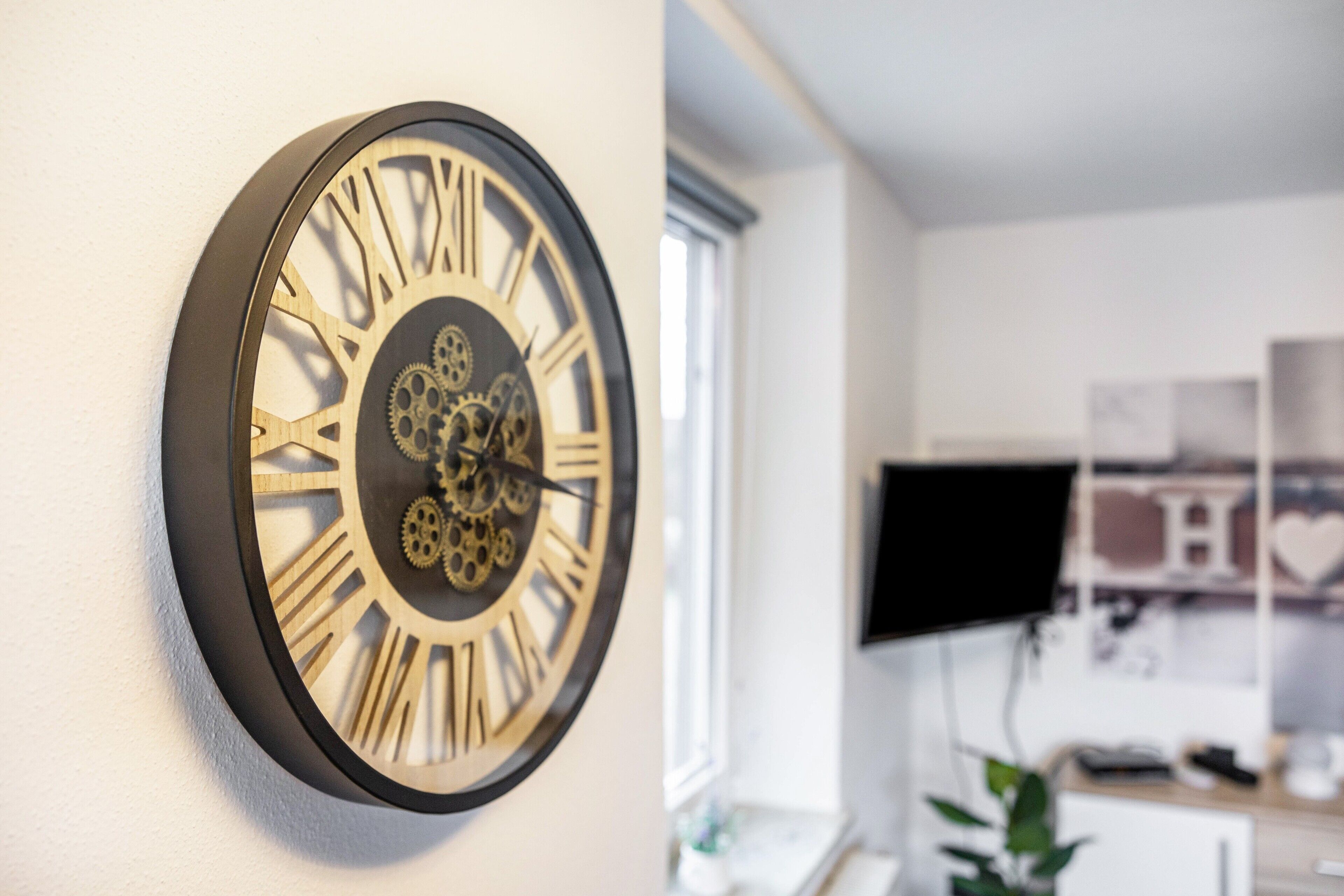 Een wandklok met Romeinse cijfers en tandwielen, een raam, en een bureau met een computer monitor.