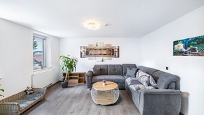 Un salon moderne avec un canapé d’angle gris, une table basse ronde, un banc en osier et une plante en pot.