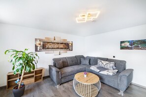 Ein modernes Wohnzimmer mit einem grauen Ecksofa, einem runden Holztisch, einer Topfpflanze und einem wandmontierten Regal.