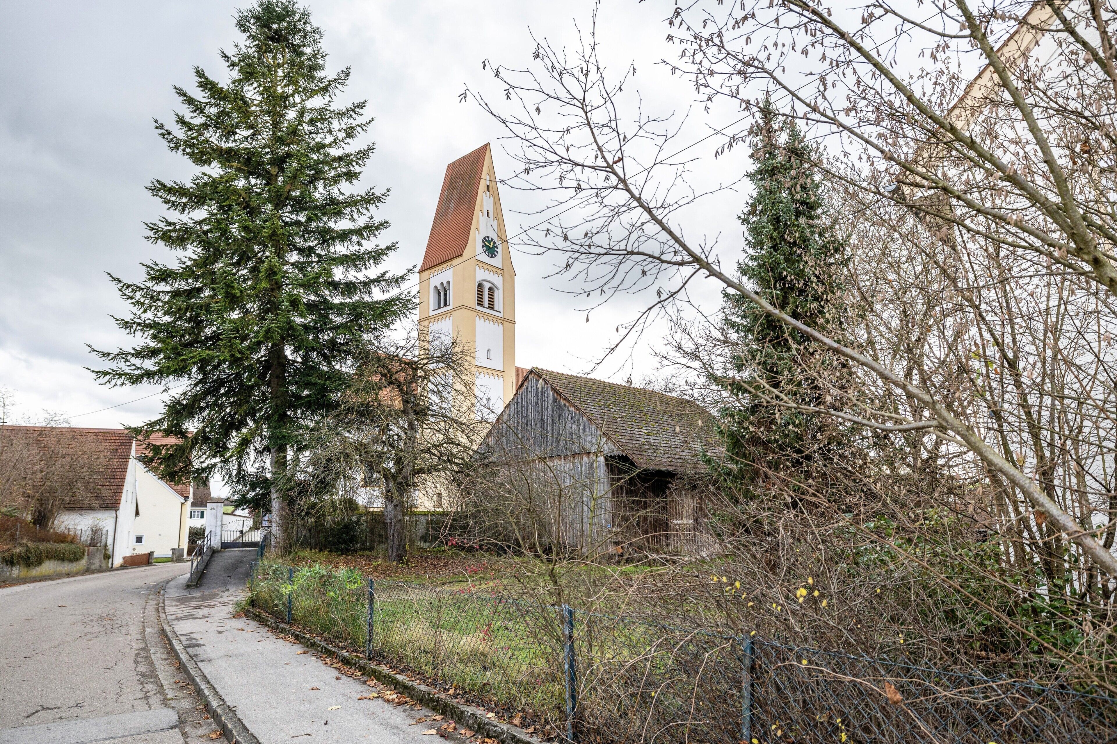 Une église dotée d’un clocher, un espace clos envahi par la végétation et une route menant à un bâtiment blanc.