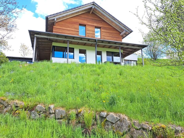 Una casa moderna de madera con techo inclinado, ventanas amplias y un muro de contención de piedra.