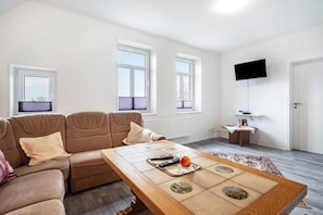 Ein Wohnzimmer mit einer Couch, einem Couchtisch mit Fernbedienungen und Obst, einem Fernseher an der Wand und einer Tür.