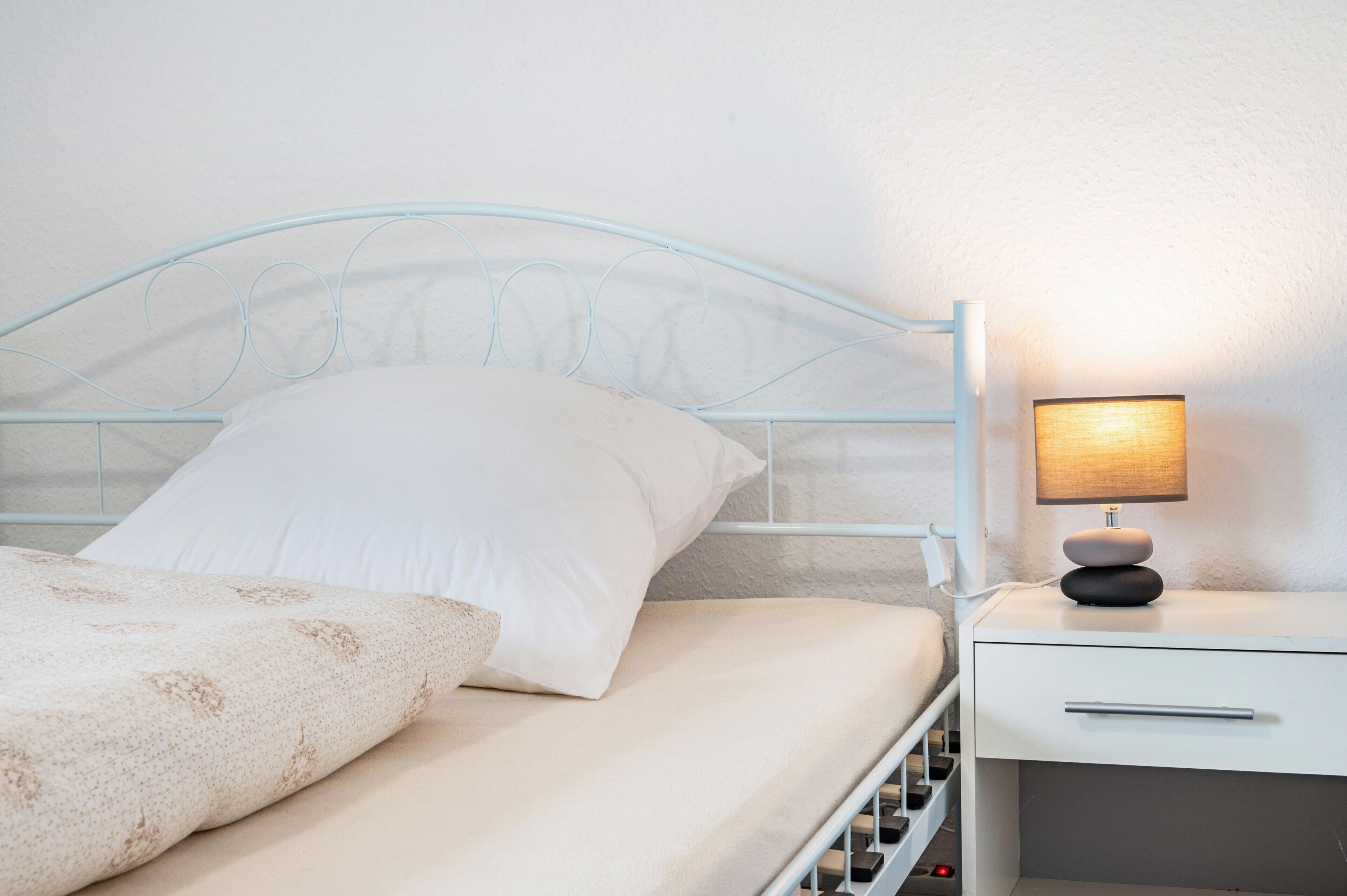A bed with a white headboard, a bedside table with a lamp, and a white drawer.