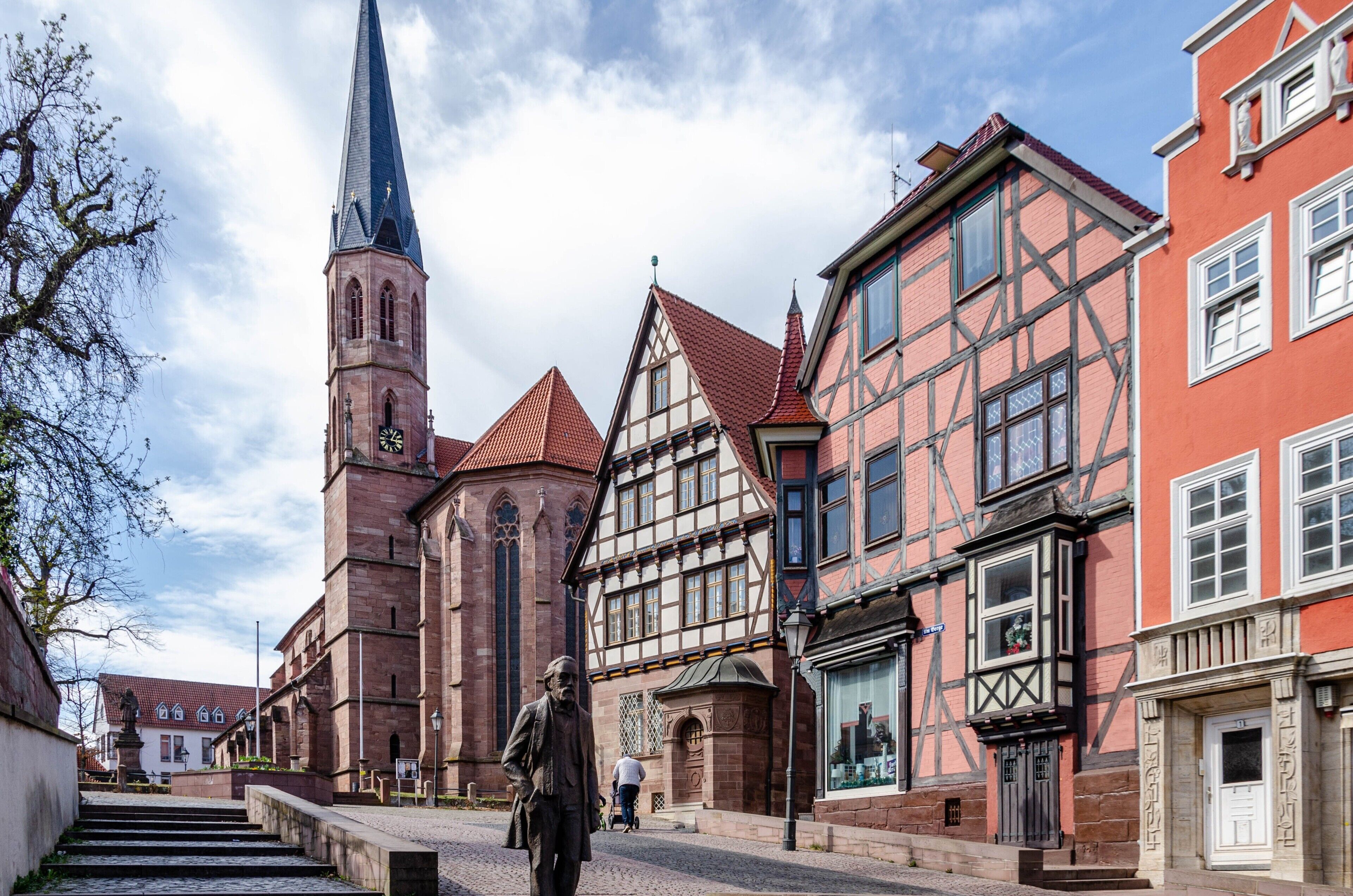 A historic building with a prominent tower, adjacent half-timbered houses, and a statue in the foreground.